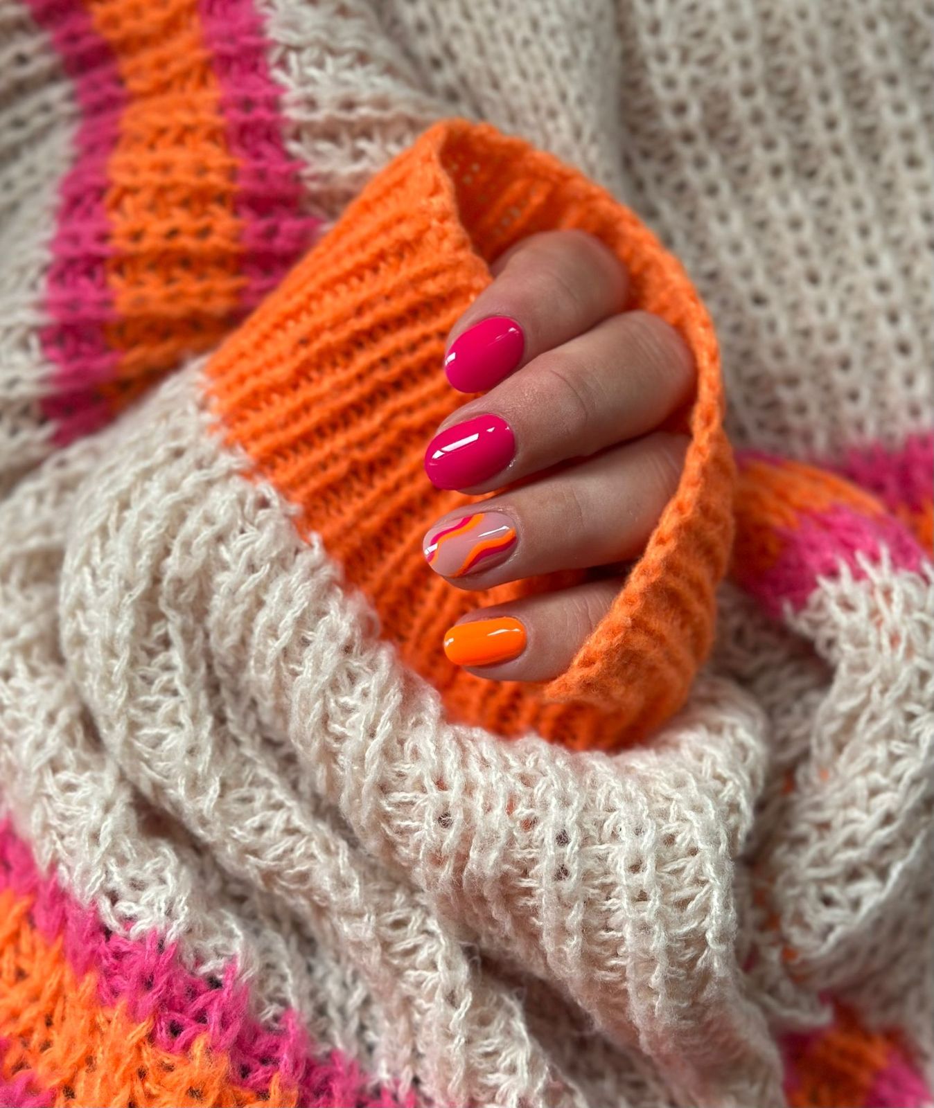 Korte nagels in fuchsia, roze en oranje met een streepjesdesign, in een gebreide trui